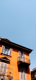 A vibrant yellow-orange building features two floors with wooden shutters and balconies adorned with ornate railings. Small plants add a touch of greenery at the balcony edges. The sky is clear and bright, contributing to a warm and inviting atmosphere.