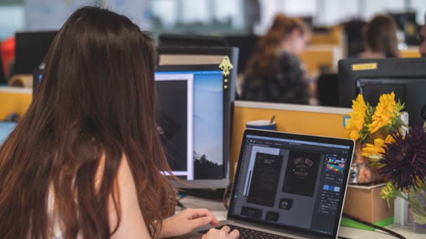 A person with long hair is working at a computer in a modern office environment. The desk is equipped with multiple monitors, and a laptop is displaying design software. Bright yellow and purple flowers are placed on the desk, adding a cheerful touch. Other workers can be seen in the background, focused on their tasks.