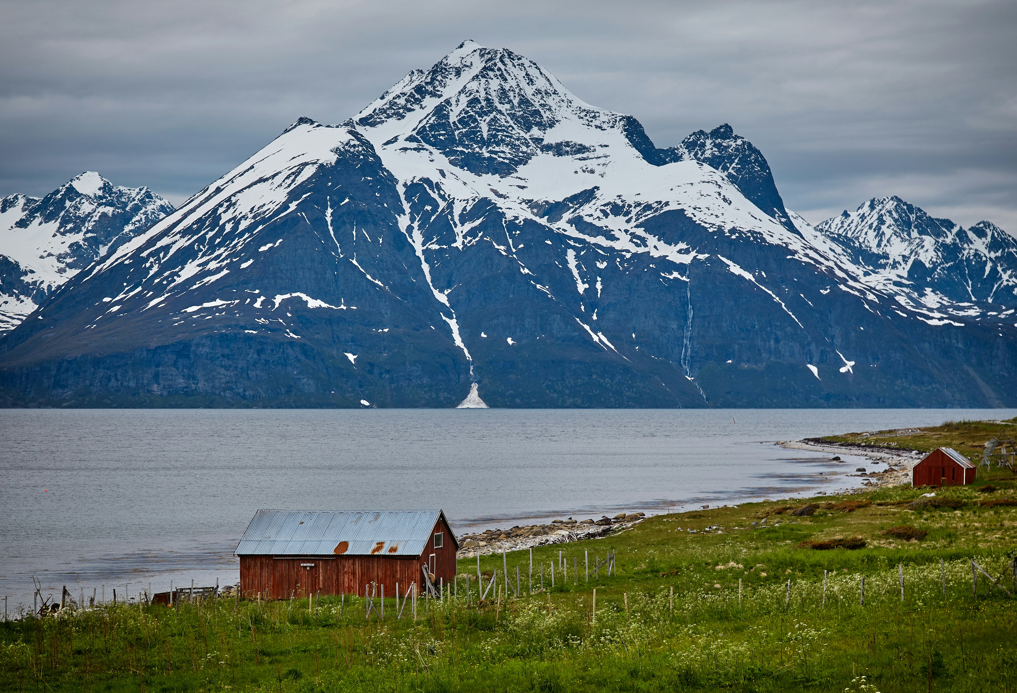 photo of two brown wooden barns near blue body of water and snow covered mountains at daytime
