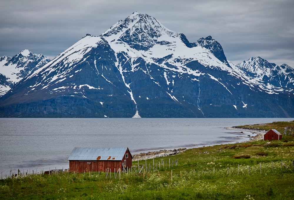 photo of two brown wooden barns near blue body of water and snow covered mountains at daytime