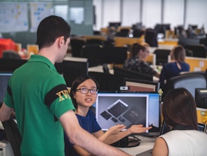 A group of people are in an office setting, engaged in discussion around a computer screen displaying design mockups. The atmosphere suggests a collaborative work environment with attention focused on the digital content.