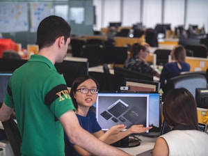 A group of people are in an office setting, engaged in discussion around a computer screen displaying design mockups. The atmosphere suggests a collaborative work environment with attention focused on the digital content.