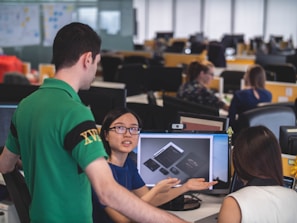 A group of people are in an office setting, engaged in discussion around a computer screen displaying design mockups. The atmosphere suggests a collaborative work environment with attention focused on the digital content.