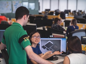 A group of people are in an office setting, engaged in discussion around a computer screen displaying design mockups. The atmosphere suggests a collaborative work environment with attention focused on the digital content.