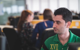 A man wearing a green polo shirt is seated in an office environment. He is looking intently in one direction, possibly focused or deep in thought. In the background, two women with blurred faces are engaged in conversation or work, sitting at desks with computers. The setting appears to be a modern open-plan office with a soft, natural light coming through large windows.