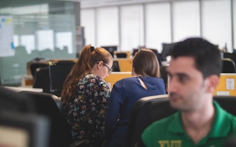 Several people are working in a modern open-plan office with computers on desks. Two people are sitting closely, engaged in conversation or collaboration. The environment appears busy, with a focus on work.
