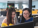 Two women are focused on computer screens in an open office environment. The workspace is bright and modern, with partitions, multiple desks, and visible computer monitors. In the foreground, there are yellow flowers, adding a touch of color to the scene.