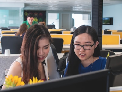 Two women are focused on computer screens in an open office environment. The workspace is bright and modern, with partitions, multiple desks, and visible computer monitors. In the foreground, there are yellow flowers, adding a touch of color to the scene.