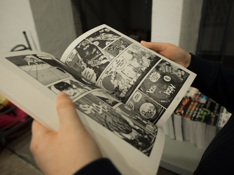 A person is holding a black and white comic book with illustrations and text. The page displays multiple panels depicting an action scene, possibly involving dialogue and sound effects. In the background, a stack of similar books can be seen, suggesting a bookstore or library setting.