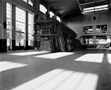 A large industrial space with high ceilings and tall, expansive windows allowing light to stream in. The room contains a massive industrial machine with pipes and ladders, suggesting a factory or power plant. The architecture is grand and symmetrical, emphasizing the scale and functionality of the space.