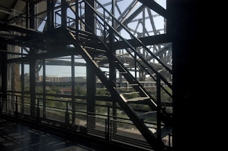 A metal staircase with a grid pattern silhouette against a backdrop of a river and cityscape. The sunlight casts shadows through the structure, highlighting the geometric lines of the architecture.