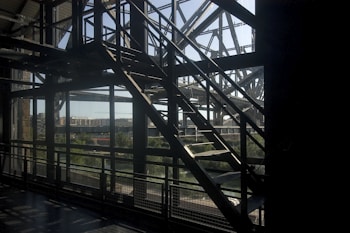 A metal staircase with a grid pattern silhouette against a backdrop of a river and cityscape. The sunlight casts shadows through the structure, highlighting the geometric lines of the architecture.