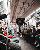 A person is performing an acrobatic move in the aisle of a subway train, surrounded by seated passengers. The train's interior is detailed with steel bars and overhead lights. Passengers display varying expressions of surprise and amusement.
