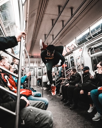 A person is performing an acrobatic move in the aisle of a subway train, surrounded by seated passengers. The train's interior is detailed with steel bars and overhead lights. Passengers display varying expressions of surprise and amusement.
