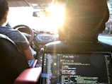 Inside a car, a person is driving with a visible steering wheel and dashboard. The setting sun casts a warm glow through the windshield. In the foreground, a laptop on the backseat displays code on its screen, suggesting someone is programming or working on a project while traveling.