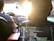 Inside a car, a person is driving with a visible steering wheel and dashboard. The setting sun casts a warm glow through the windshield. In the foreground, a laptop on the backseat displays code on its screen, suggesting someone is programming or working on a project while traveling.