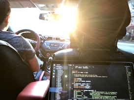 Inside a car, a person is driving with a visible steering wheel and dashboard. The setting sun casts a warm glow through the windshield. In the foreground, a laptop on the backseat displays code on its screen, suggesting someone is programming or working on a project while traveling.