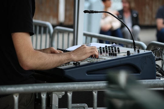 A person operates an audio mixing console, adjusting the controls with their hands. The mixer is positioned behind metal barriers, suggesting an outdoor event or concert setting. In the background, there are blurred images of people seated, possibly audience members or event staff.
