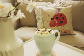A cozy living space featuring a decorative pillow with a playful ladybug design and heart motifs, accompanied by the text 'kisses'. In the foreground is a light blue mug filled with a whipped topping. A neutral-toned couch with patterned pillows and a white table carrying a vase with white flowers complete the scene.