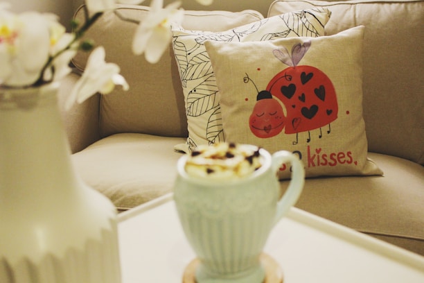 A cozy living space featuring a decorative pillow with a playful ladybug design and heart motifs, accompanied by the text 'kisses'. In the foreground is a light blue mug filled with a whipped topping. A neutral-toned couch with patterned pillows and a white table carrying a vase with white flowers complete the scene.