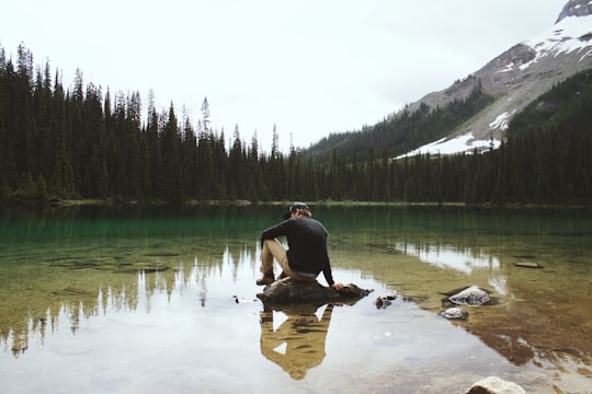 man wearing sweater sitting on stone by calm lake with trees on shore fronting snow capped mountain at daytime in Yoho National Park Of Canada Canada