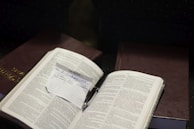 An open book with text is placed on top of another closed book. A small handwritten note is inserted between the pages of the open book, with text referencing 'II Corinthians 10:5'. A pen is resting near the note.