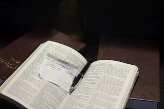 An open book with text is placed on top of another closed book. A small handwritten note is inserted between the pages of the open book, with text referencing 'II Corinthians 10:5'. A pen is resting near the note.