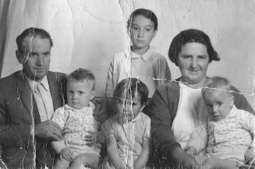 A vintage black-and-white photograph depicting a family gathered closely. An adult man in a suit and a woman with short hair are seated. Between them and in front of them are four small children. The image shows signs of wear with visible creases and marks, suggesting it is quite old.