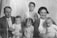 A vintage black-and-white photograph depicting a family gathered closely. An adult man in a suit and a woman with short hair are seated. Between them and in front of them are four small children. The image shows signs of wear with visible creases and marks, suggesting it is quite old.
