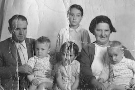 A vintage black-and-white photograph depicting a family gathered closely. An adult man in a suit and a woman with short hair are seated. Between them and in front of them are four small children. The image shows signs of wear with visible creases and marks, suggesting it is quite old.