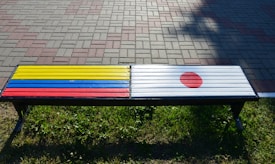 A bench is painted with the colors and symbols of two national flags. The left side displays horizontal stripes in yellow, blue, and red, while the right side features a white background with a red circle. The bench is situated on a grassy area adjacent to a paved walkway composed of multicolored bricks.
