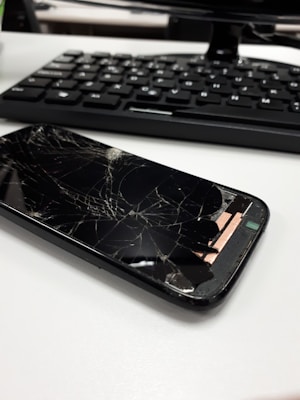 A smartphone with a severely cracked screen is lying on a white surface. In the background, there is a keyboard, suggesting an office or work environment.