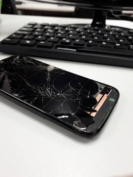 A smartphone with a severely cracked screen is lying on a white surface. In the background, there is a keyboard, suggesting an office or work environment.