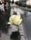 A white rose is inserted into the engraved name on a reflective black stone memorial. In the background, an American flag is in focus, with people observing along the edge. Trees and tall buildings are visible, creating a solemn atmosphere.