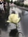 A white rose is inserted into the engraved name on a reflective black stone memorial. In the background, an American flag is in focus, with people observing along the edge. Trees and tall buildings are visible, creating a solemn atmosphere.