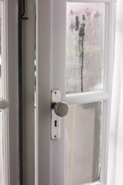 An interior view of a white wooden door with glass panels, featuring a classic round doorknob and a keyhole. The glass is foggy with visible streaks of moisture, suggesting condensation or rain. Sunlight filters through, illuminating the area with a gentle glow.