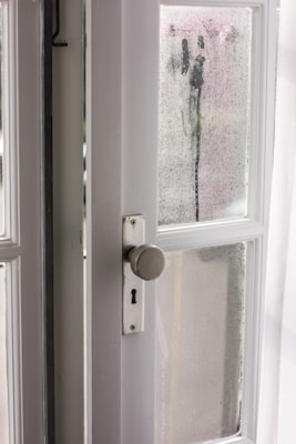 An interior view of a white wooden door with glass panels, featuring a classic round doorknob and a keyhole. The glass is foggy with visible streaks of moisture, suggesting condensation or rain. Sunlight filters through, illuminating the area with a gentle glow.