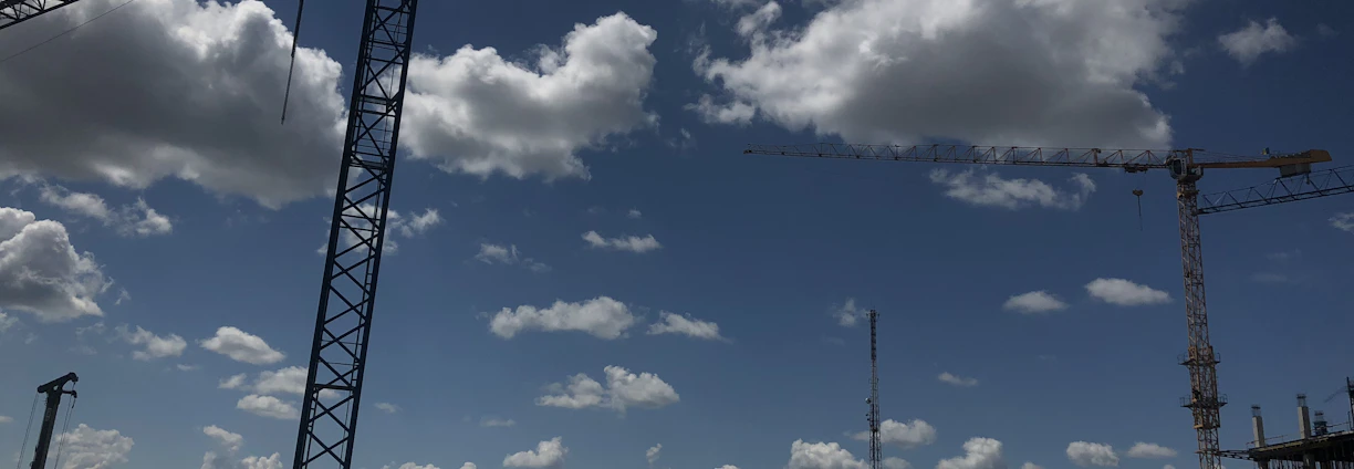 A bustling construction site with cranes lifting materials, set against a green landscape under a clear sky.