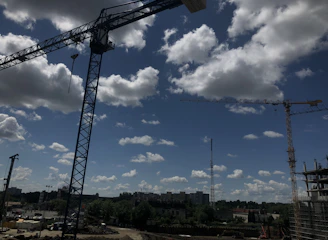 A bustling construction site with cranes lifting materials, set against a green landscape under a clear sky.