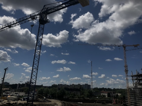 A construction site with large cranes under a partly cloudy sky. Several buildings and trees are visible in the background, and the ground is filled with machinery and construction materials.
