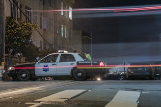 A police car is parked on a city street at night, with light trails from passing vehicles creating streaks of red and white across the scene. Nearby buildings are partially illuminated, and a suspension bridge with lights is visible in the background.