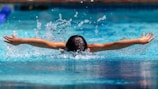 A person is swimming in a pool, doing the butterfly stroke with arms outstretched and water splashing around them. The scene captures the dynamic movement and energy of swimming.