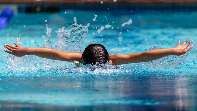 A person is swimming in a pool, doing the butterfly stroke with arms outstretched and water splashing around them. The scene captures the dynamic movement and energy of swimming.