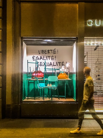 A store window display with a bold, artistic arrangement that includes stacked chairs and large handbags. The wall behind has French words painted in black: 'Liberté! Égalité! Sexualité!'. The setting suggests a blend of fashion and political messaging. A blurred individual walks past the window on the street outside.