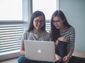Two people are sitting indoors and looking at a laptop. They both have long, dark hair and are wearing glasses. One person is wearing a light-colored t-shirt, and the other is dressed in a striped shirt. They are sitting on the floor next to each other in a bright room with large windows.