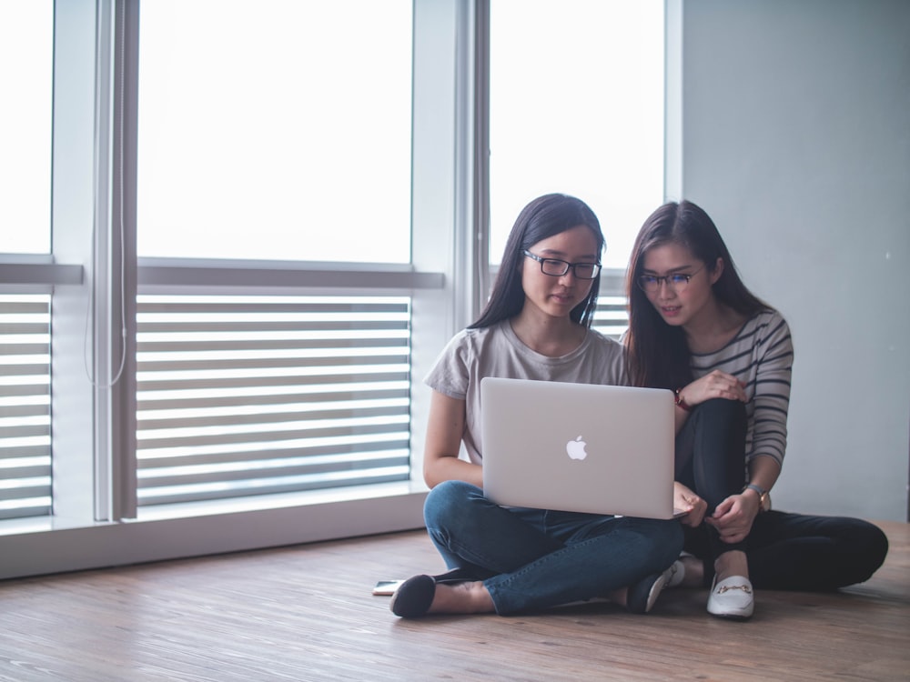 two women facing silver MacBook sitting on parquet floor