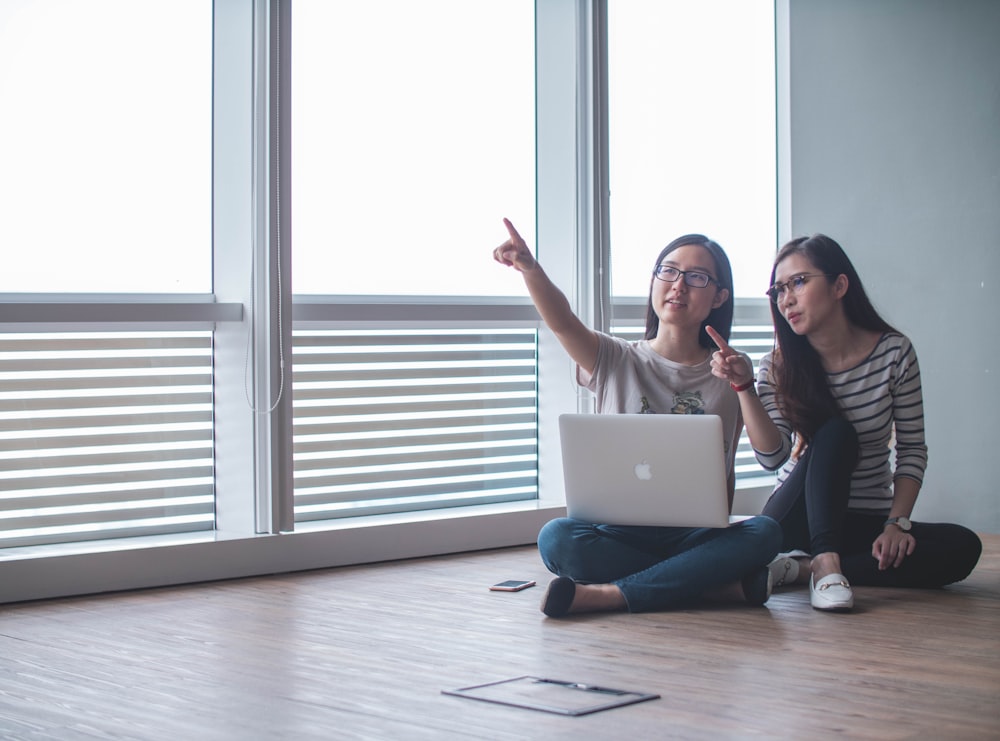 two women sitting on brown floor inside white room beside glass windows pointing upwards photo