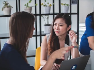 Two women are engaged in a conversation at a table in an office setting. One woman gestures with her hand while the other listens attentively. A laptop with a visible logo is on the table between them. In the background, shelves with potted plants add a decorative touch to the modern workspace.