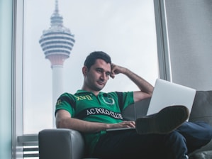 A man sitting on a couch in a relaxed posture, with a laptop on his lap. He appears to be looking at the screen with a thoughtful expression. In the background, a large tower is visible through a window, indicating an urban setting.