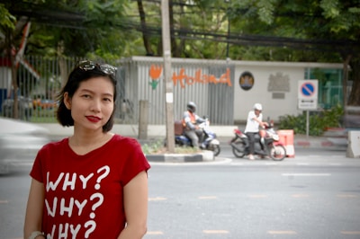 A woman wearing a red t-shirt with the text 'WHY? WHY? WHY?' is standing on a street. Behind her, two people on scooters are visible, parked near the road. There's a metal fence with the word 'Holland' written on it in orange letters and a sign indicating no parking. Trees in the background create a green, natural setting.